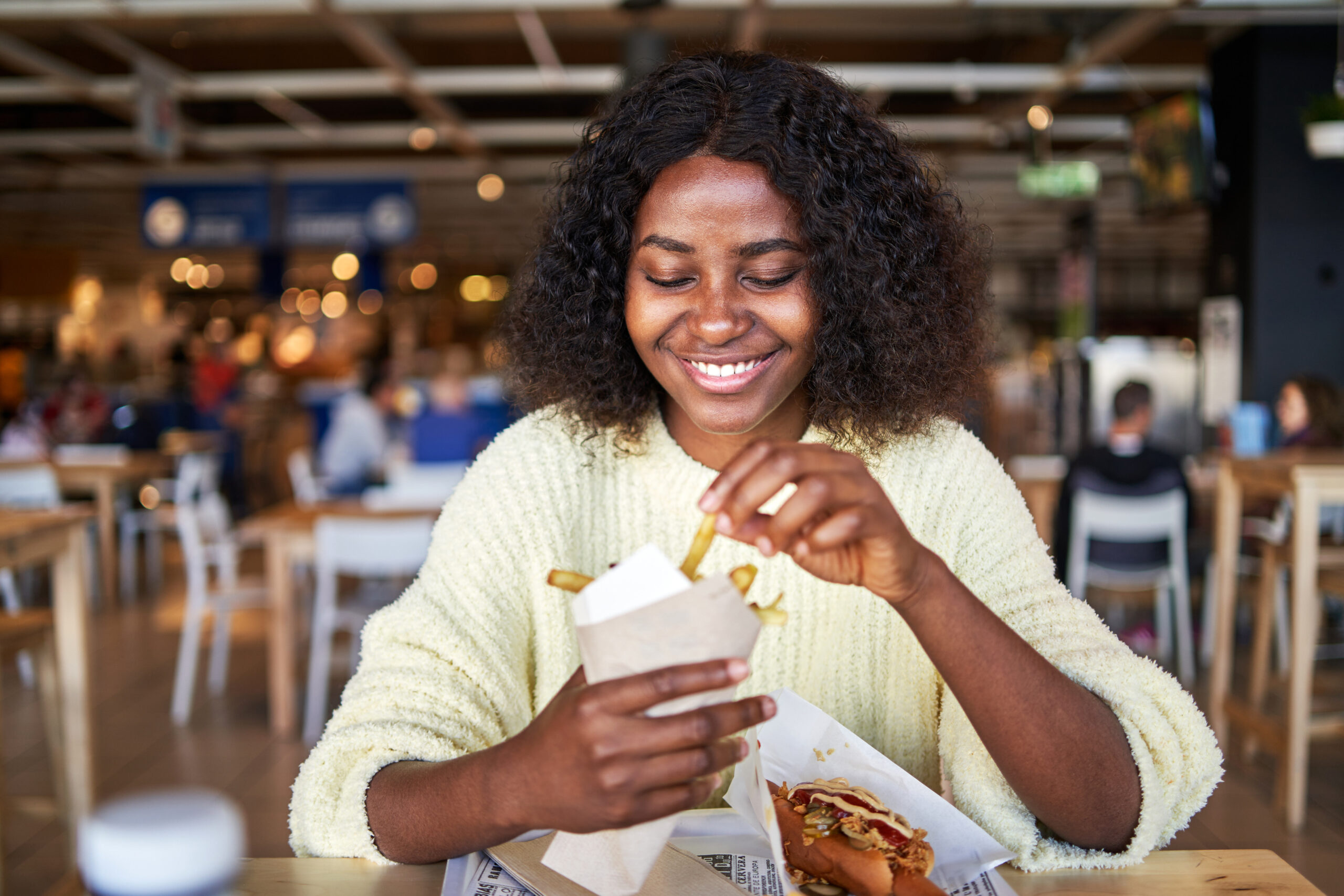 Front view of a happy young woman eating french fries and a hot dog in a shopping mall food court.