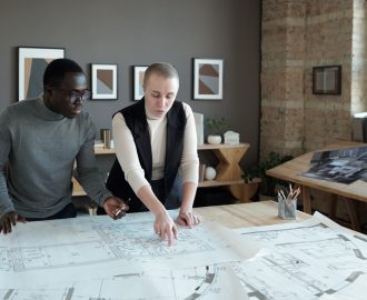 Two confident engineers in smart casualwear discussing sketch on blueprint while standing by table in large office